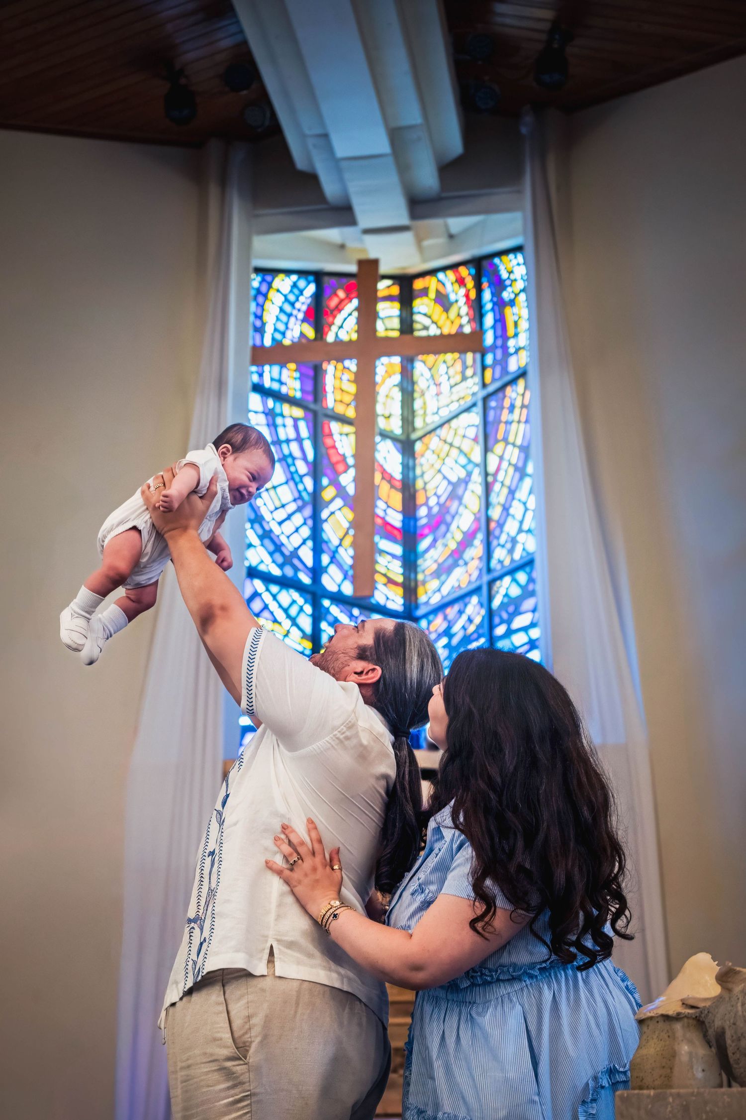 Parents lifting their baby inside a church beneath stained glass windows during a baptism celebration.