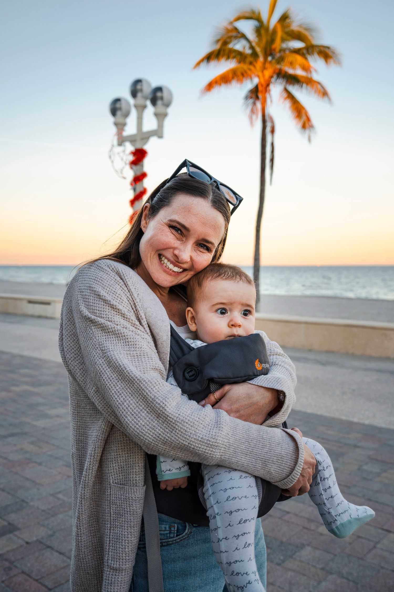 Lifestyle portrait of a woman holding a baby at sunset along the South Florida coast, captured with a warm, candid feel.