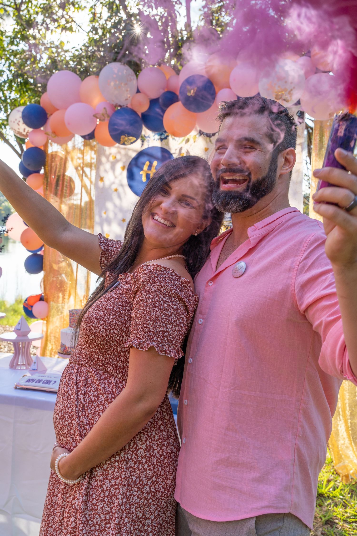 Expecting couple smiling together as pink smoke fills the air during an outdoor gender reveal in South Florida.