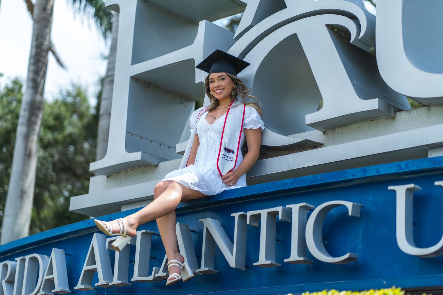 FAU graduate in white dress and cap sits confidently on FAU sign, celebrating her academic journey in South Florida.