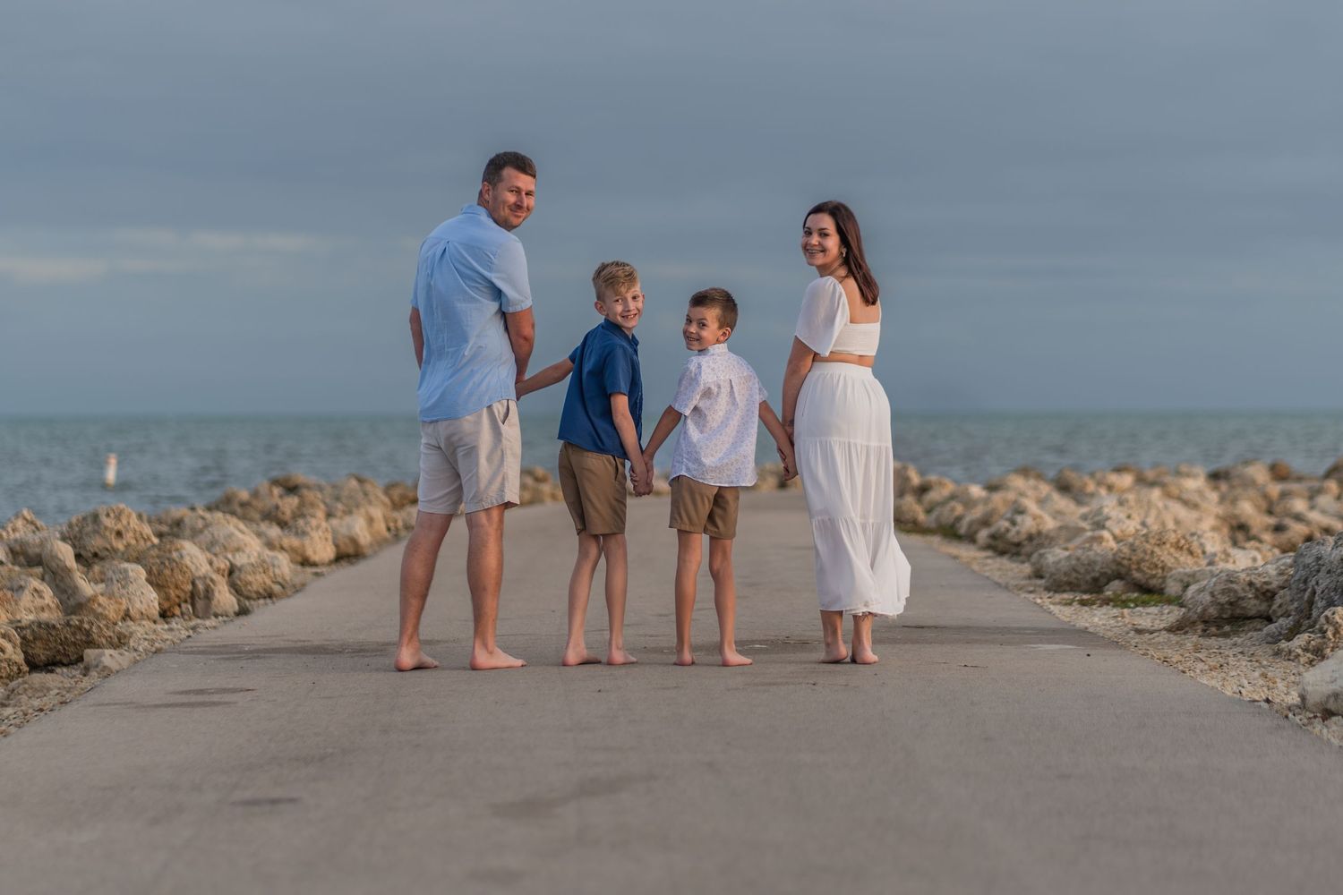 Coastal family portrait in Hollywood, FL – family walking hand in hand along a scenic rocky pier with an ocean backdrop.