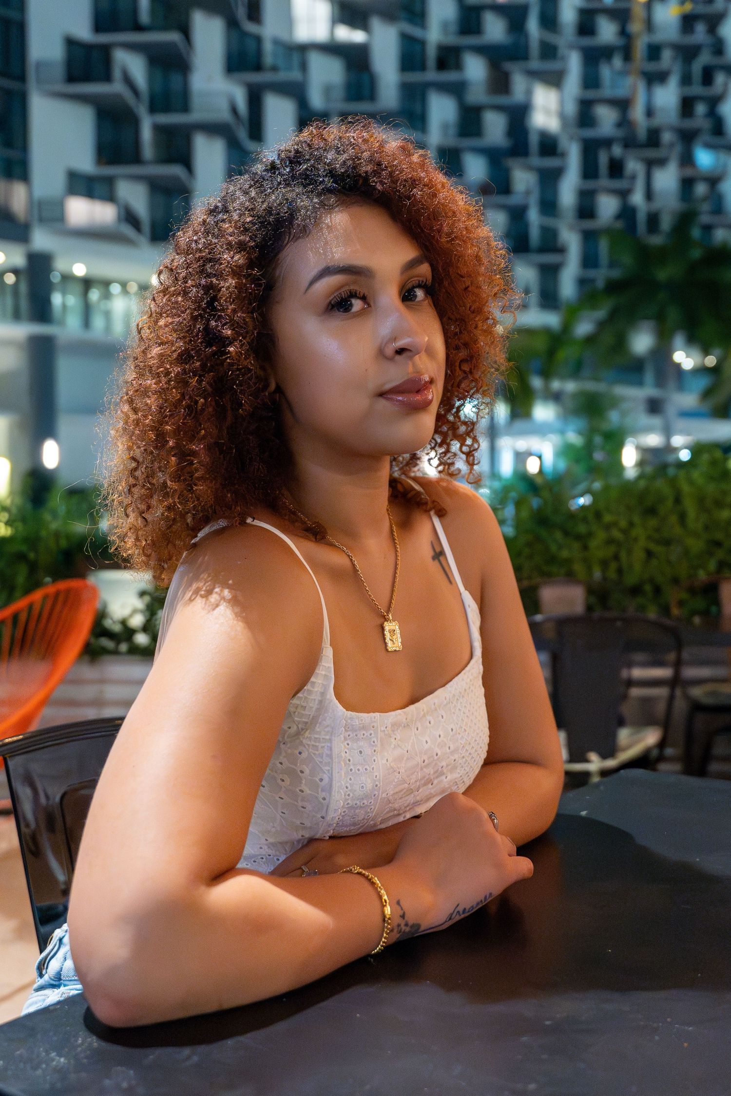 Nighttime lifestyle portrait in Brickell, Miami – curly-haired woman in a white top poses at an outdoor café with city lights behind her.