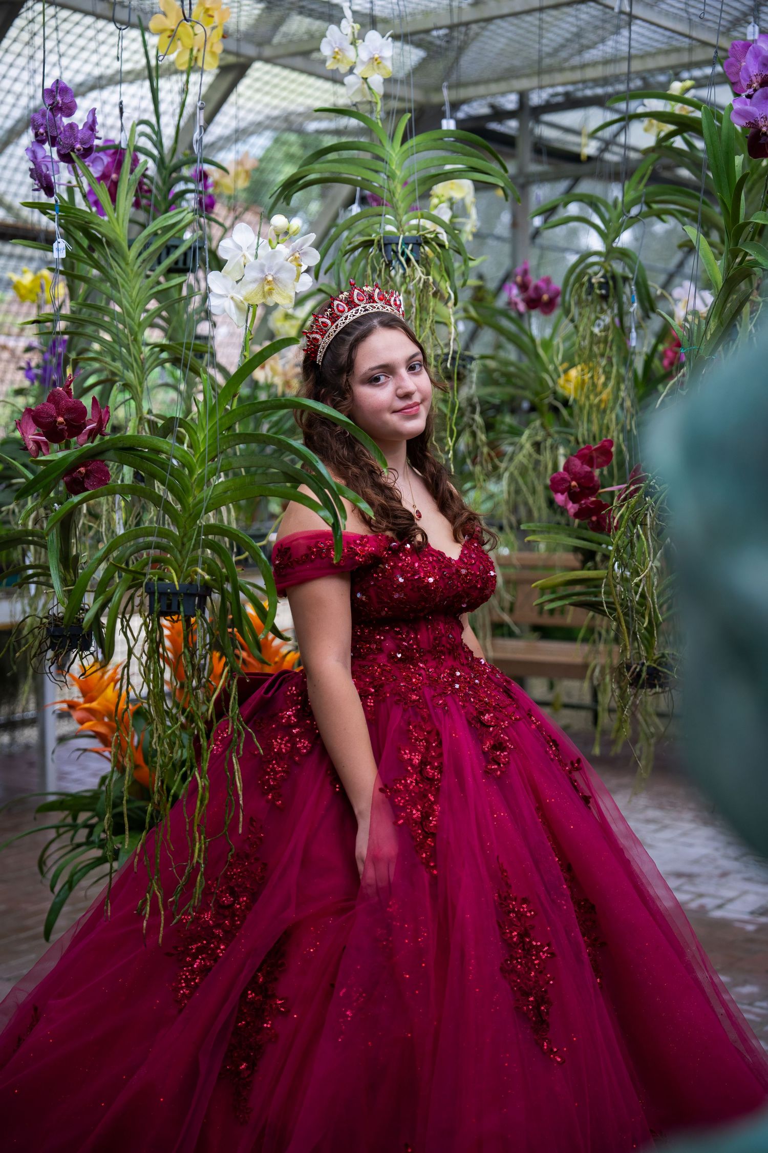 Quinceañera portrait in a burgundy gown surrounded by lush tropical plants at an orchid garden.