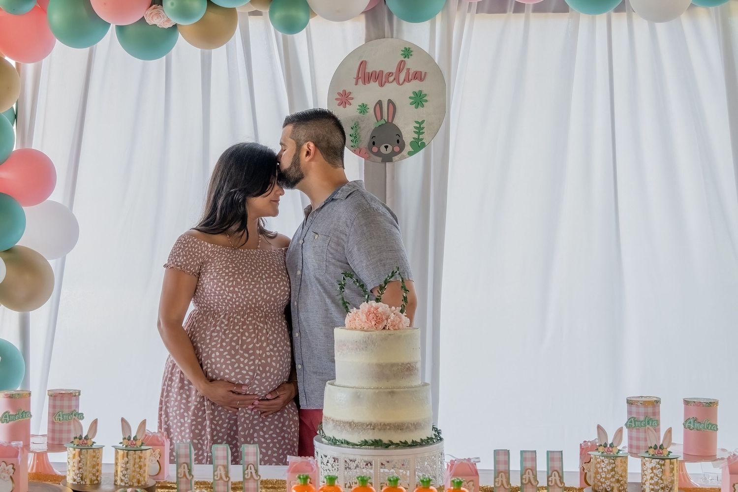 Couple sharing a kiss behind a decorated cake table during a pastel-themed baby celebration in South Florida.