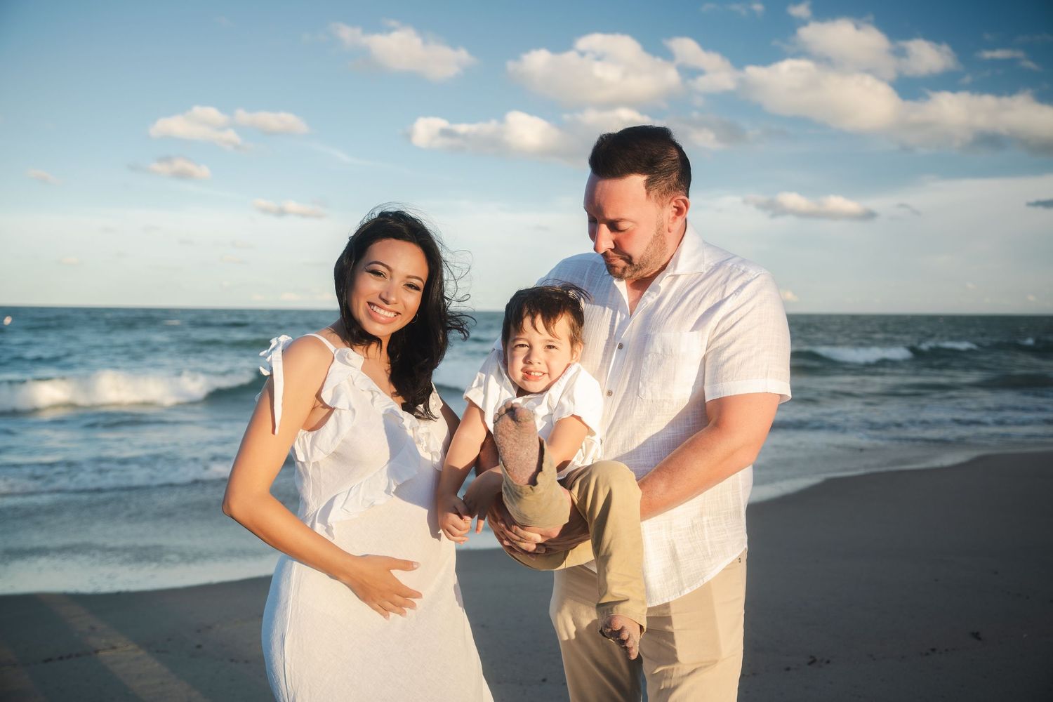 Expecting parents holding their child together on the beach during a pregnancy announcement session.