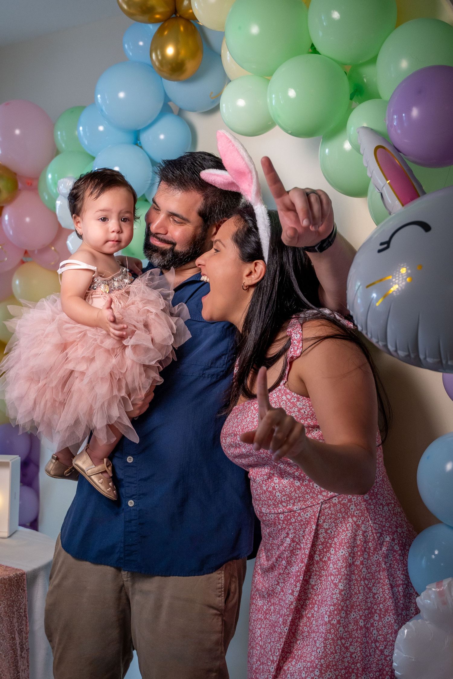 Parents holding their baby girl in front of a pastel balloon backdrop during a first birthday celebration in South Florida.