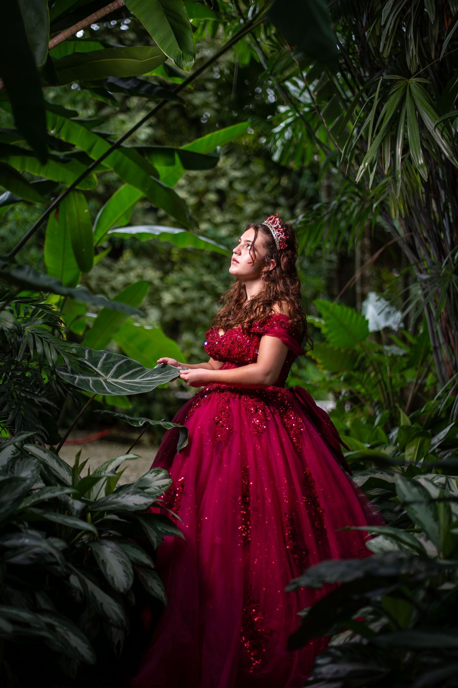 Editorial quinceañera portrait featuring a dramatic burgundy dress framed by dense greenery and soft natural light.