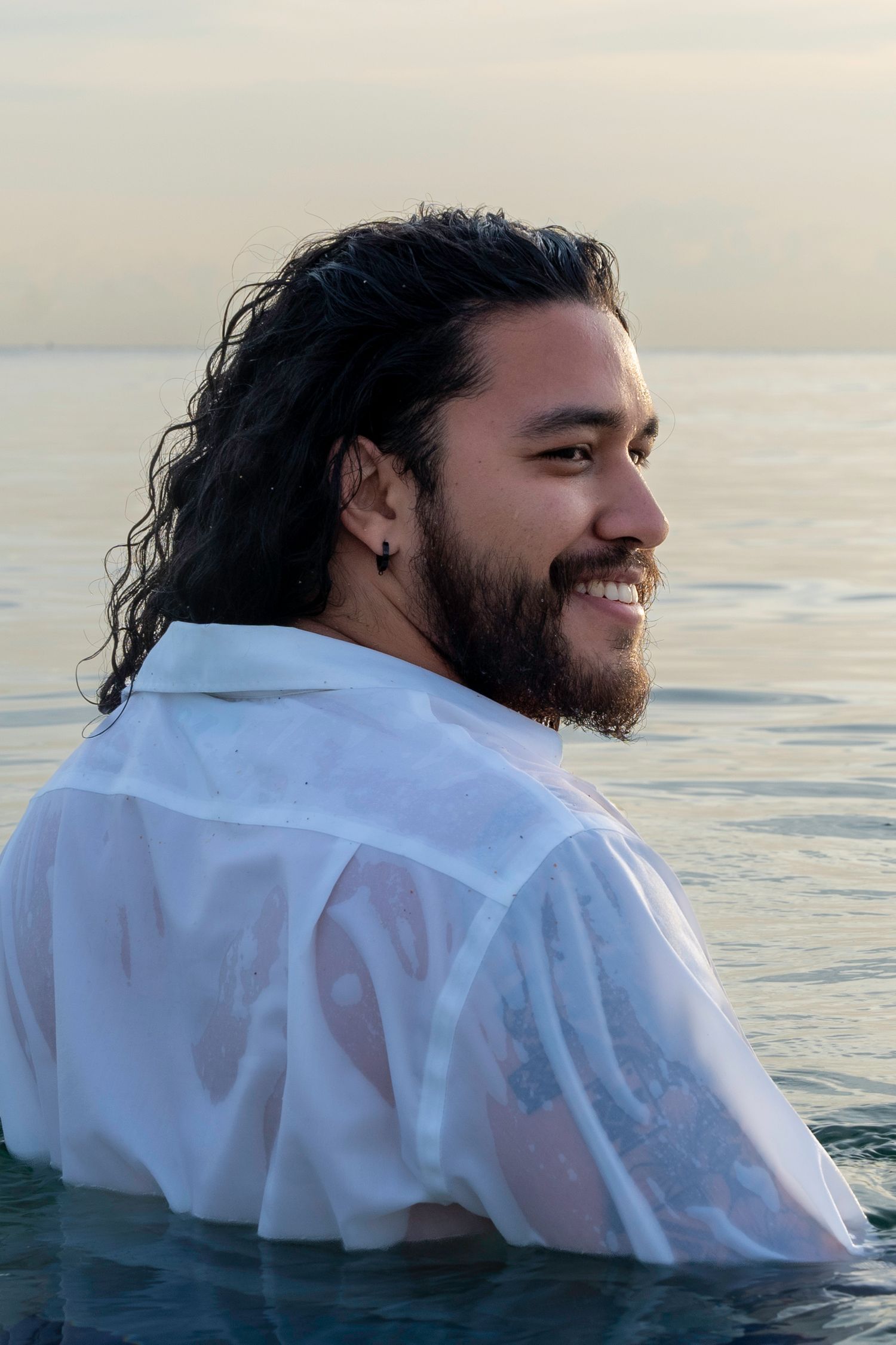 Ocean-side lifestyle photography – bearded man in a wet white shirt smiles while waist-deep in water, golden hour glow.
