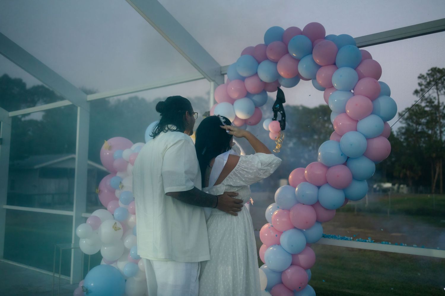 Couple embracing during a gender reveal celebration with pastel balloon decor in a South Florida backyard.