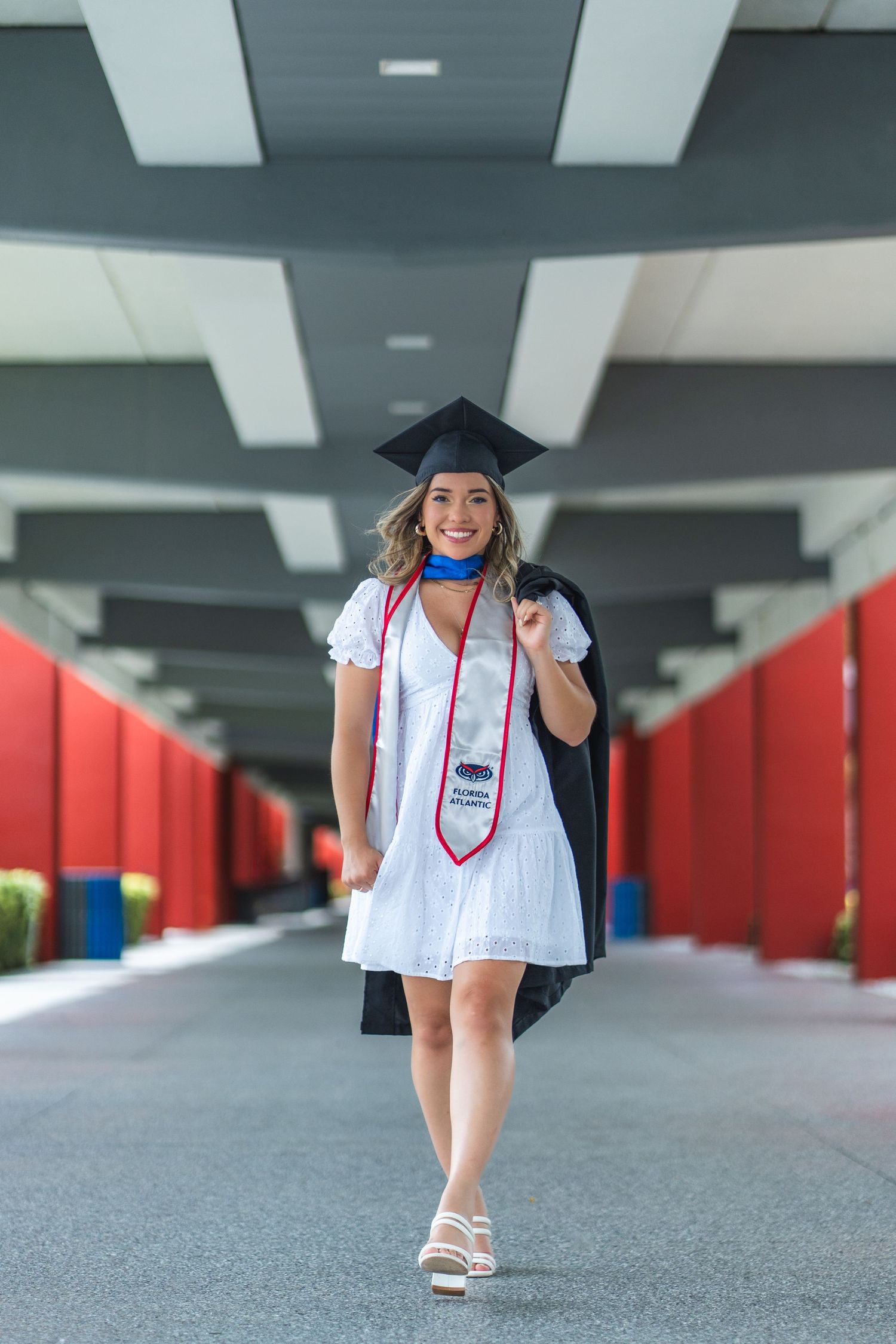 Confident FAU graduate in cap and gown walks under a modern red and black structure at Florida Atlantic University, South Florida.
