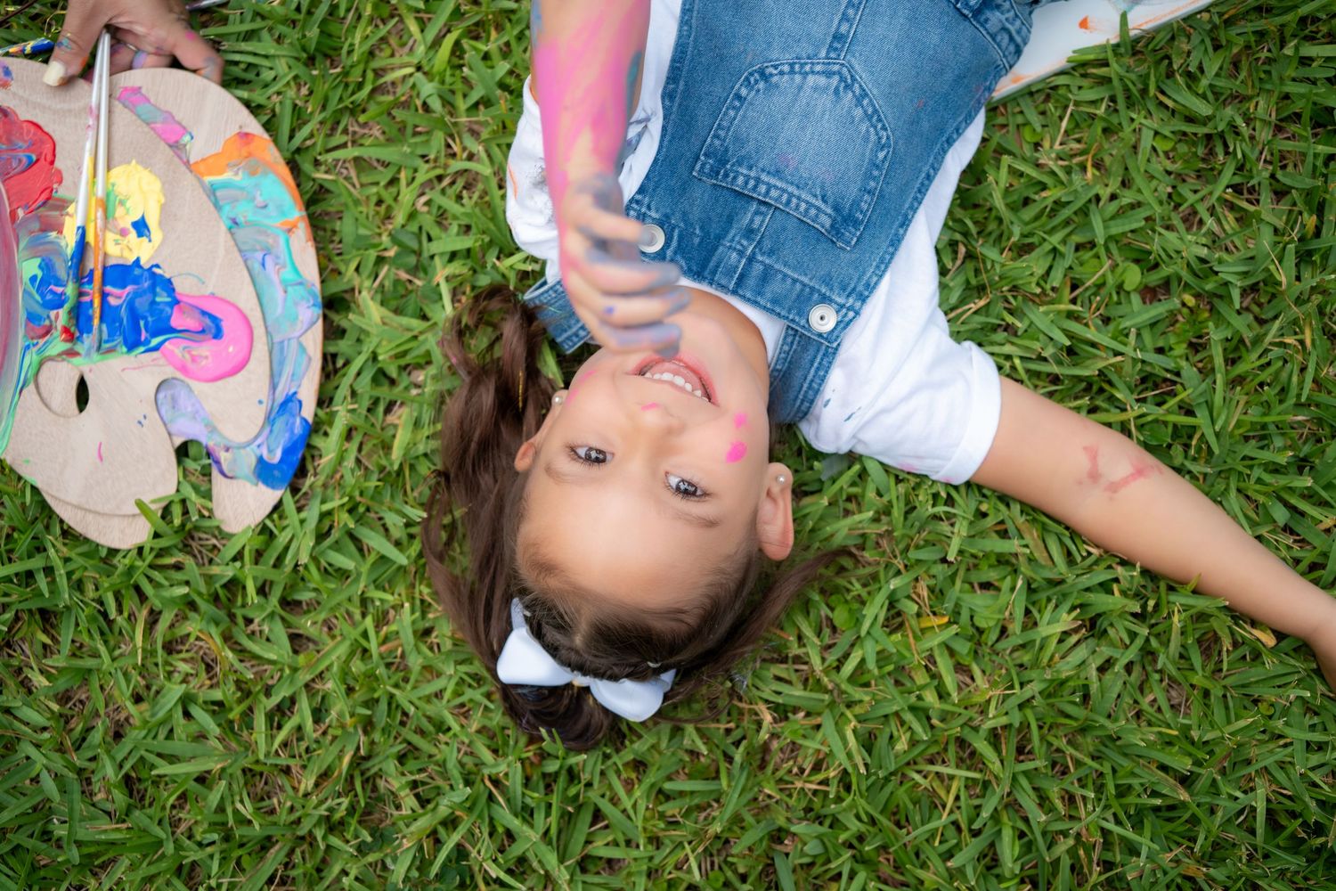 Little girl laying in the grass and smiling up at the camera with a paintbrush in hand.
