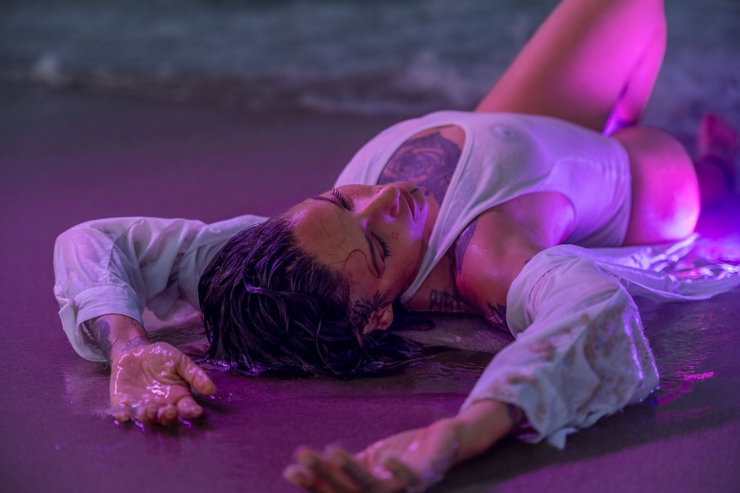 Dramatic shot of a man in a soaked white shirt, stretched across the sand under neon pink light, evoking a cinematic, dreamlike feel.