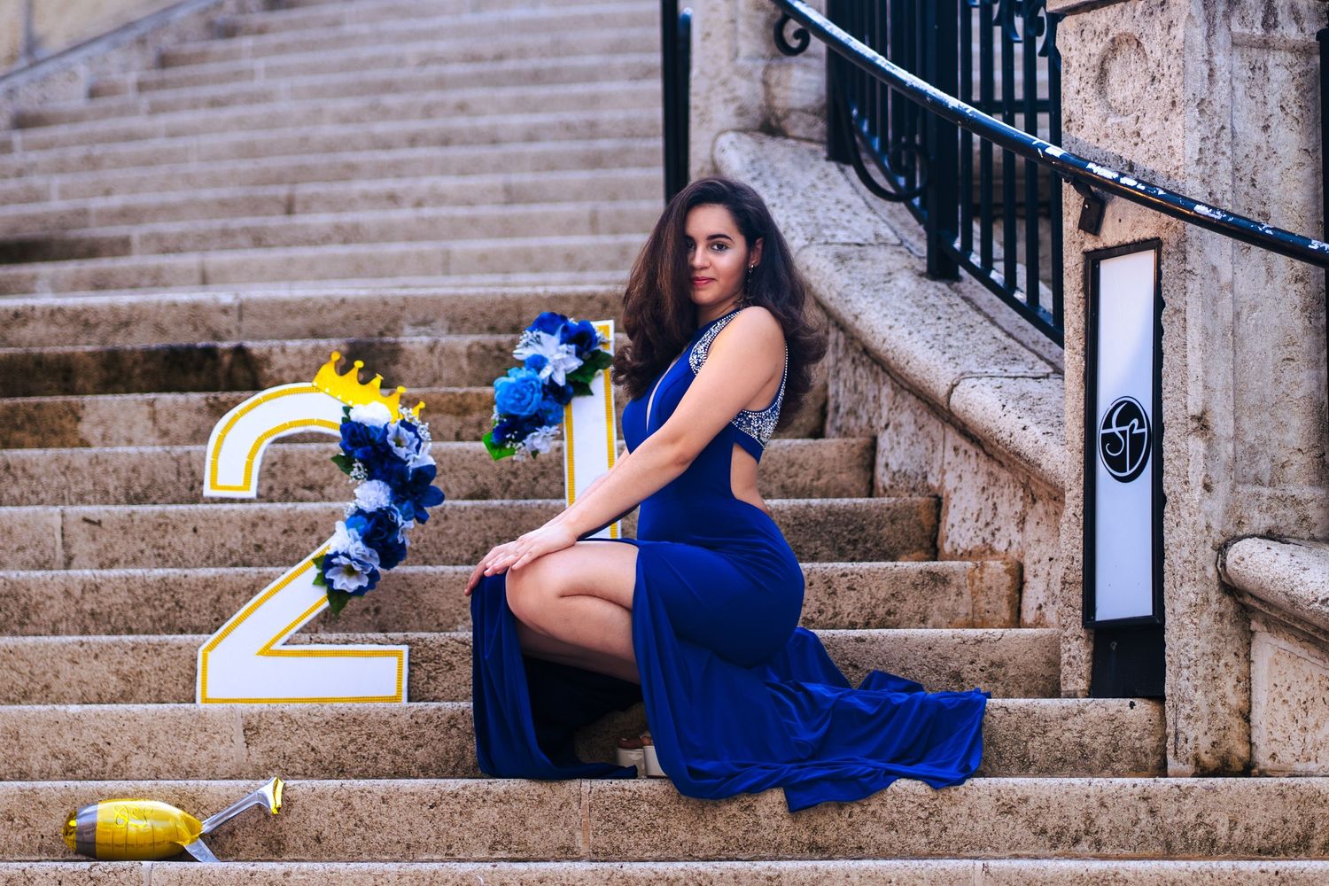 Elegant birthday portrait at Sunset Mall, in Miami FL – woman in a royal blue dress poses on stone stairs with a ‘21’ sign and flowers.