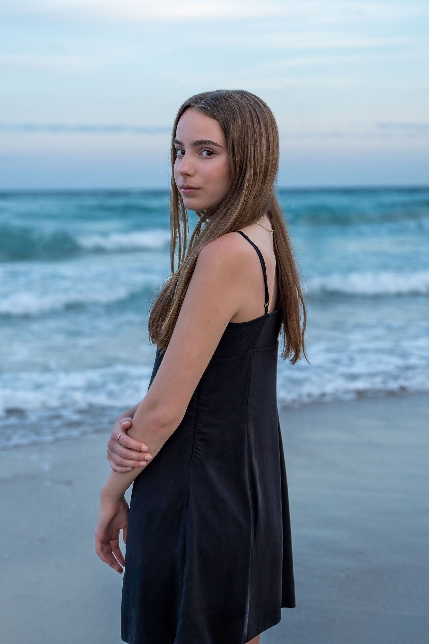A high school senior in a black dress poses against the waves, celebrating her journey with a serene ocean backdrop.