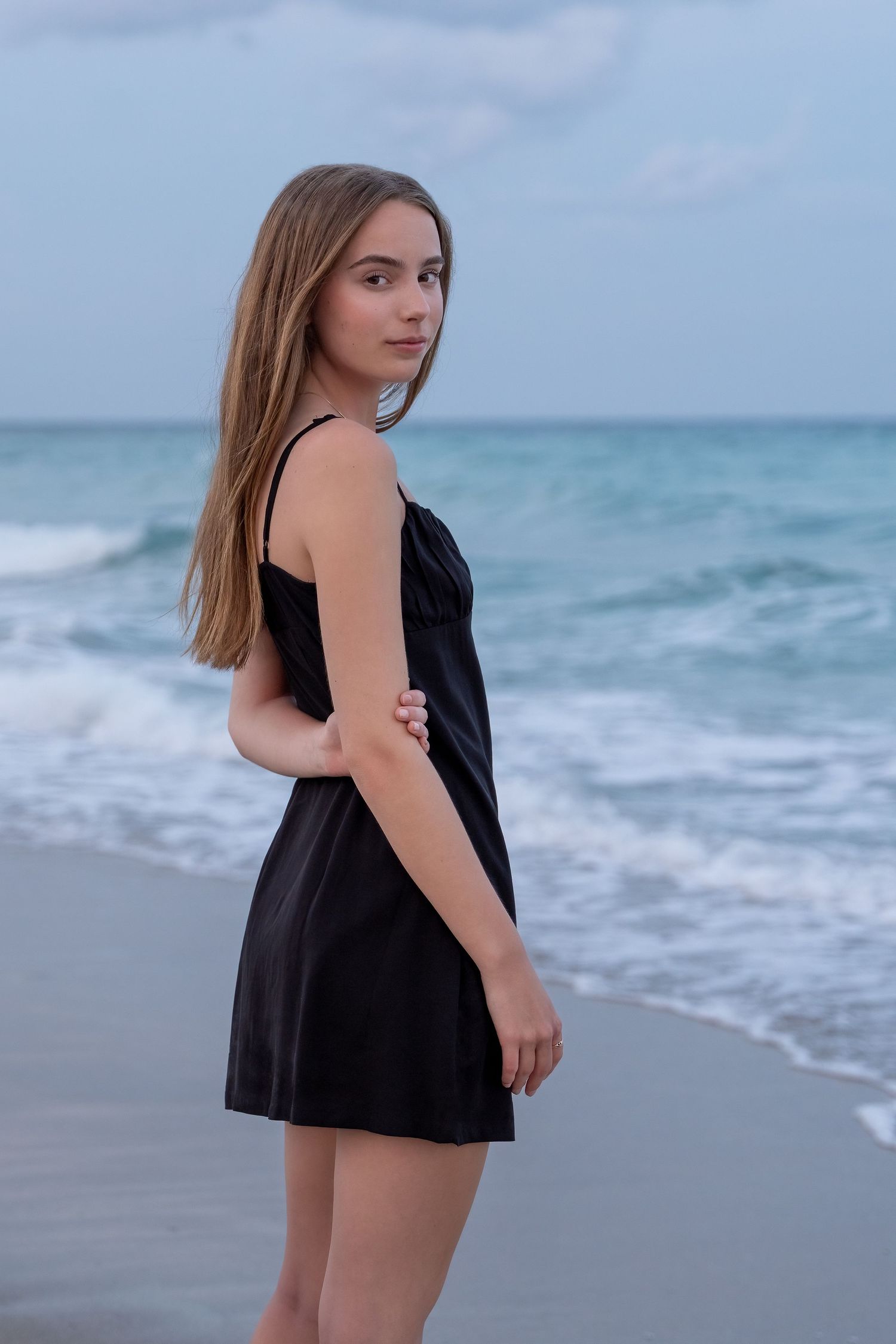 A high school senior in a black dress stands by the ocean, gazing into the distance during her beach photo session.