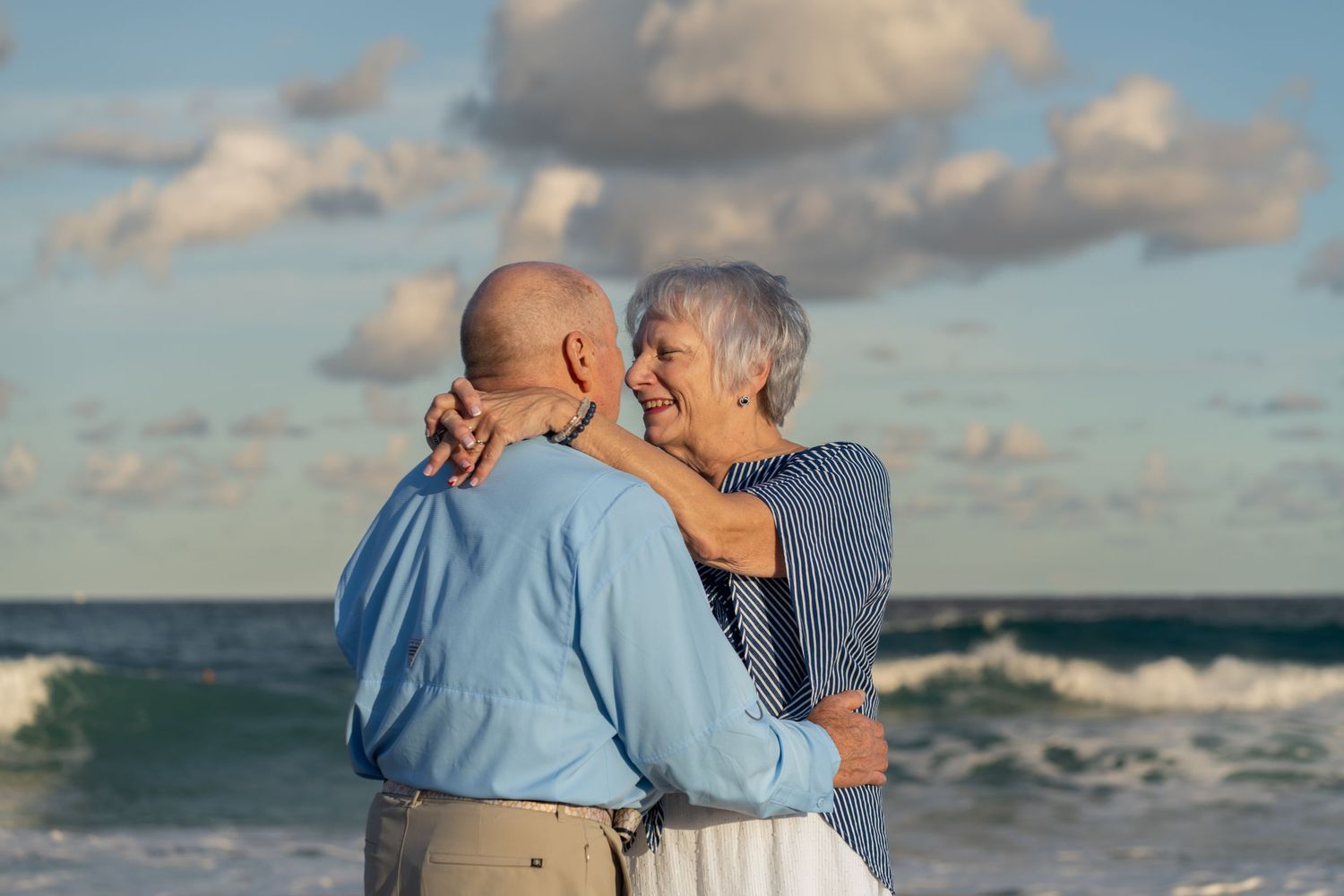 Elderly couple embracing by the ocean in Palm Beach, FL – romantic moment captured with waves crashing behind them.