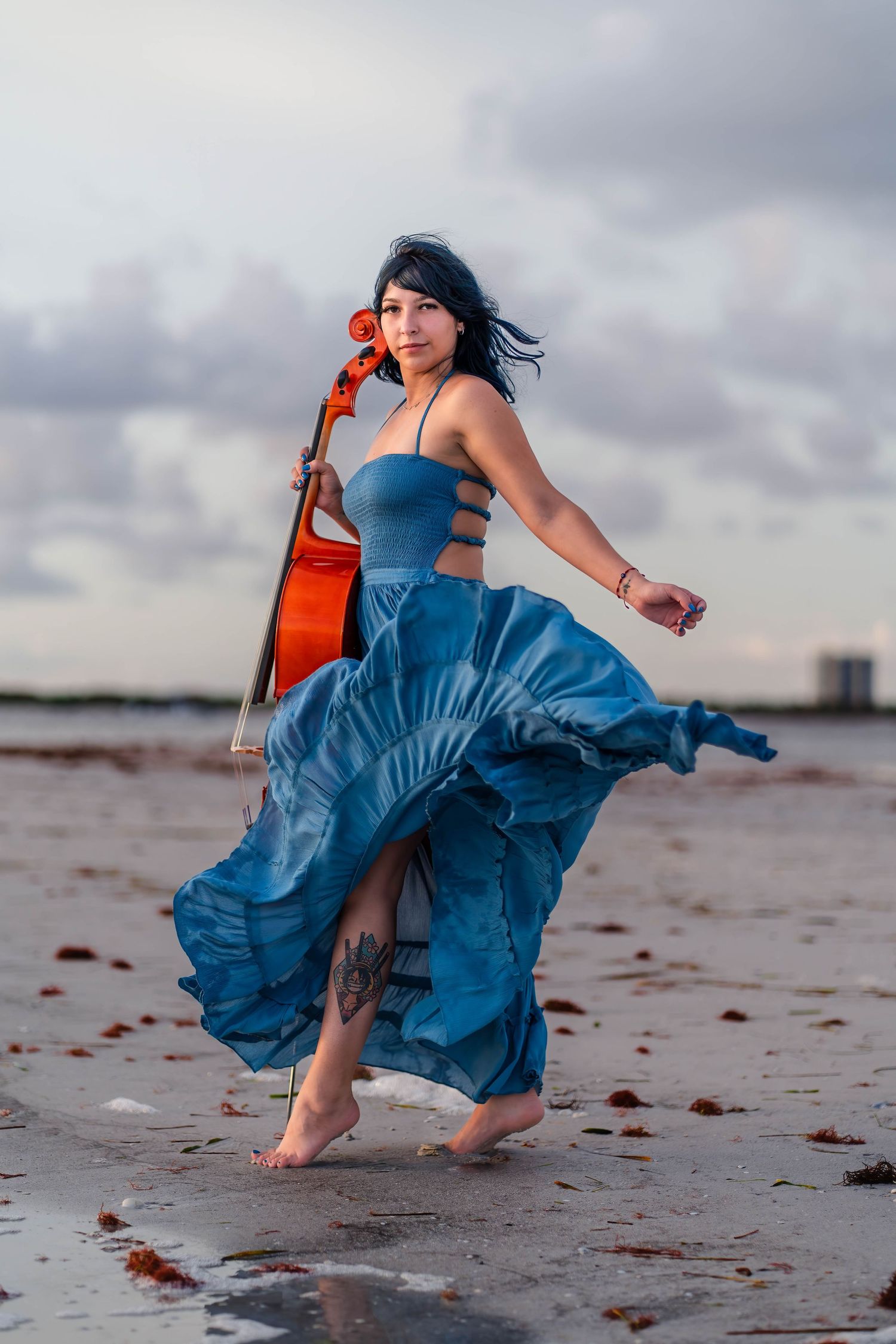 Dramatic beach portrait in Naples – woman in a flowing blue dress dances on the shore with a cello, embracing the stormy sky.