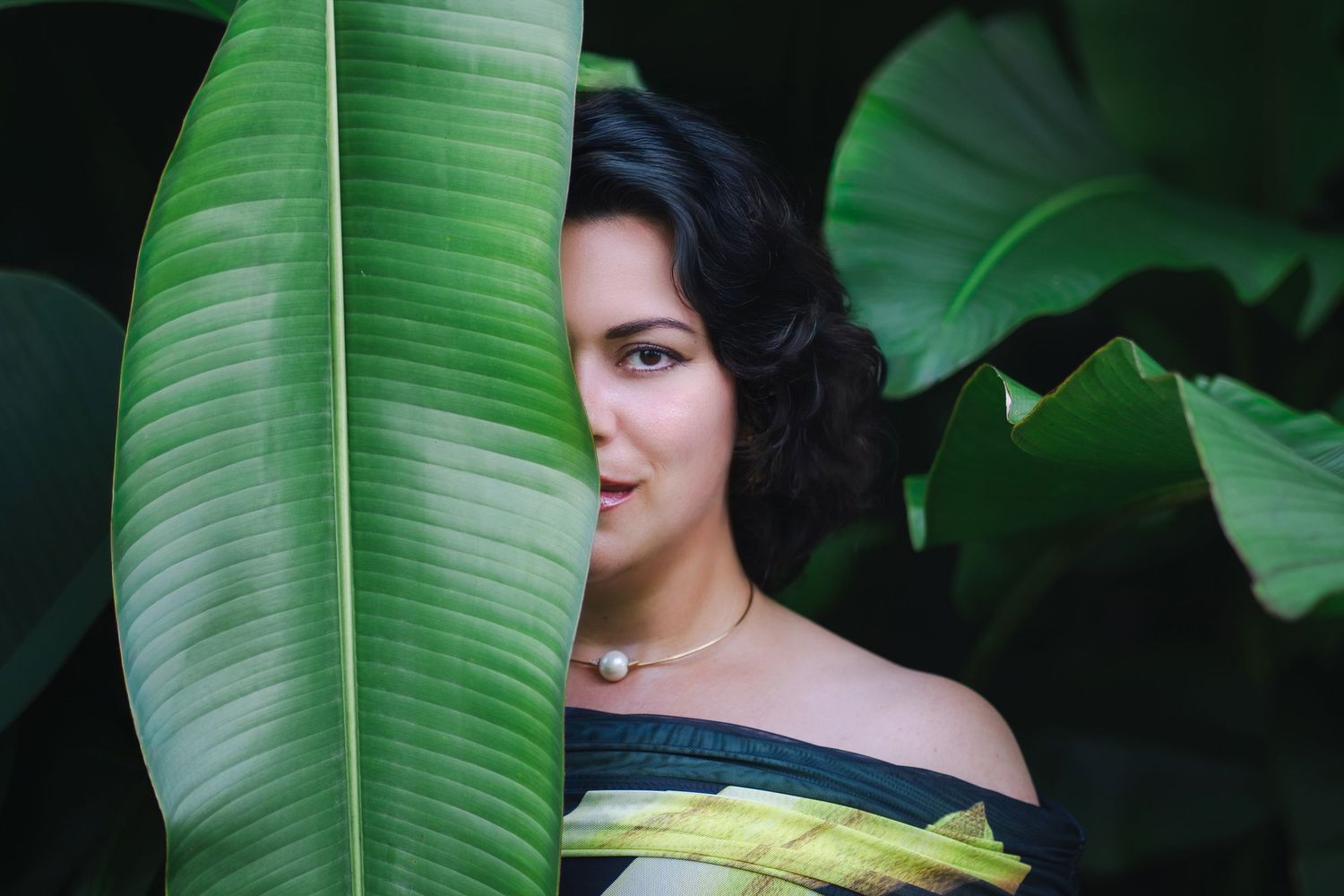 A figure poses among large tropical banana leaves in a dark moody setting with dramatic lighting.