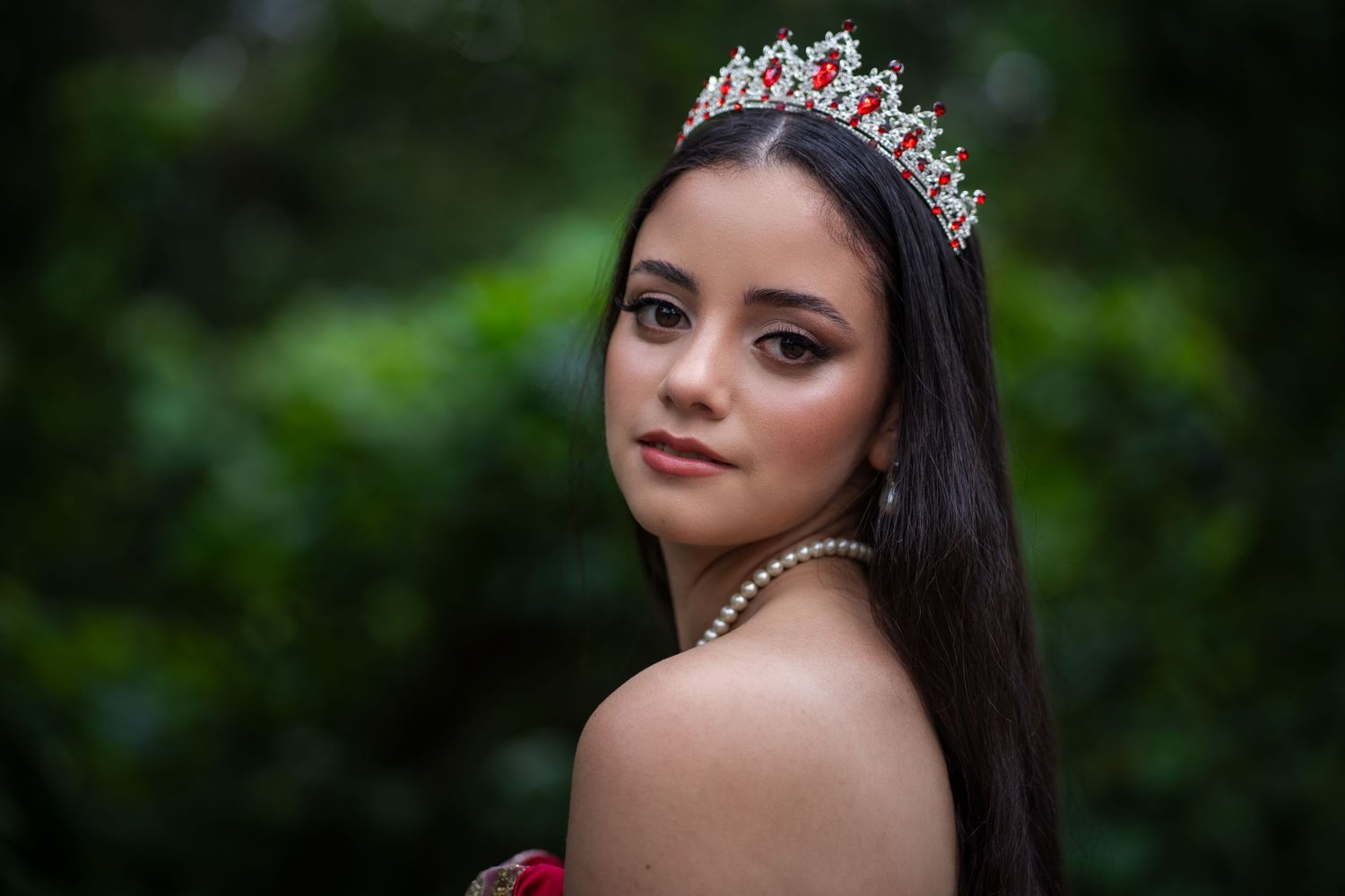 Quinceañera portrait of a girl wearing a crown and pearls, standing in front of lush greenery.