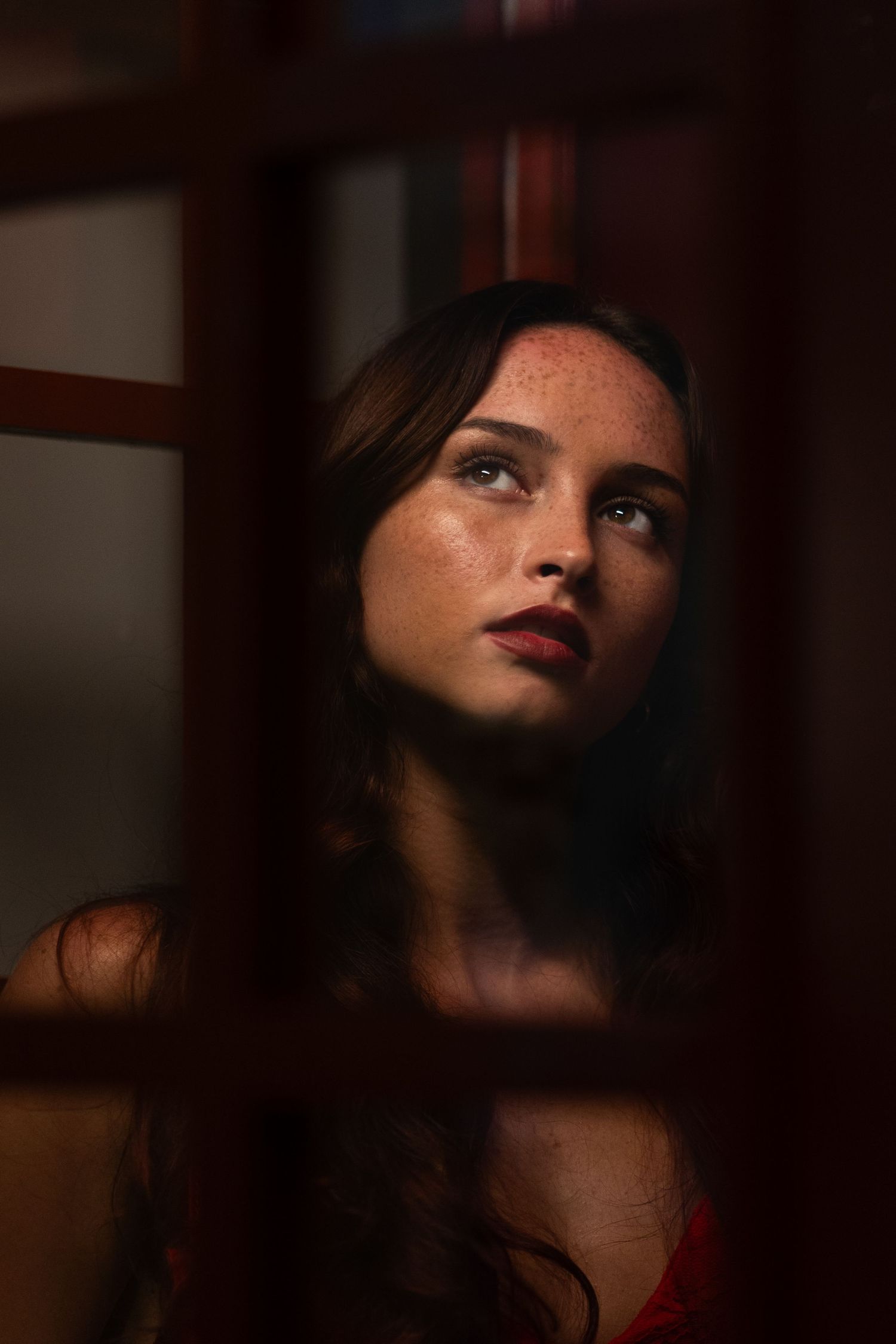Moody editorial portrait at  HGabs studio – female model with wavy brown hair gazes through a red-framed window, creating a cinematic look.