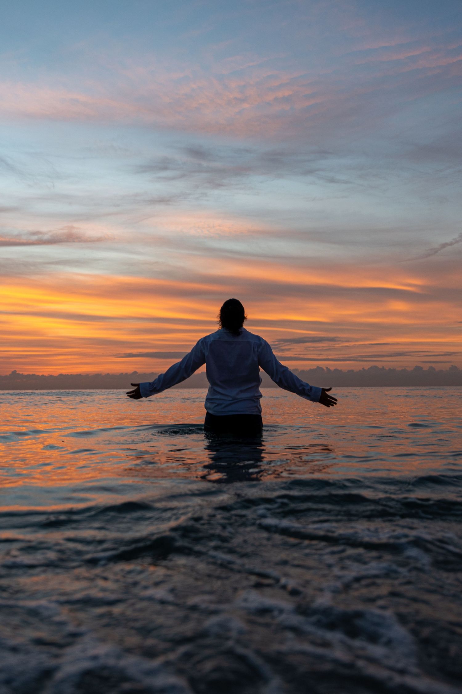 Man in a white shirt wades into the ocean at sunrise, arms outstretched toward the sky. Tranquil and cinematic beach photography.