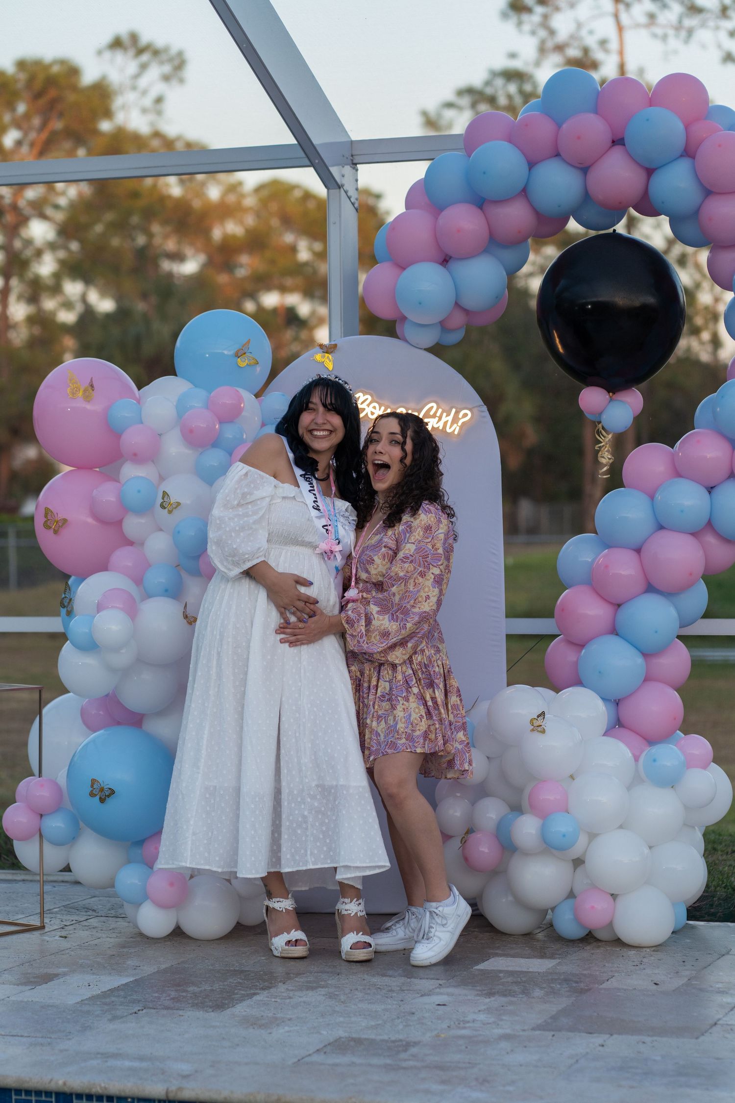 Best friends posing together beneath a pink and blue balloon arch during a South Florida gender reveal celebration.