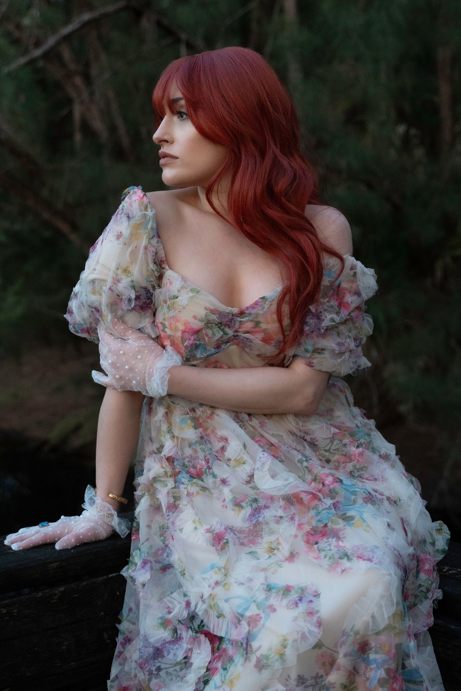 Ethereal red-haired woman in a vintage floral dress sits on a wooden log, bathed in soft golden hour light.