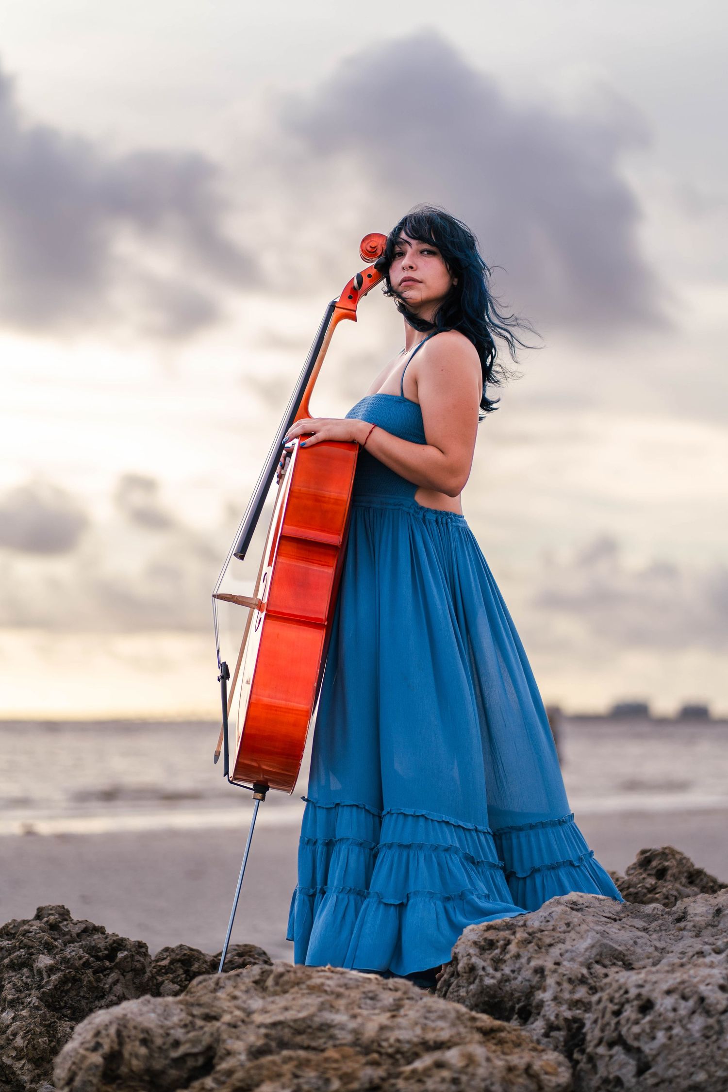 Artistic beachside cello session – woman with dark hair embraces her instrument while standing on rocky shores under moody skies.