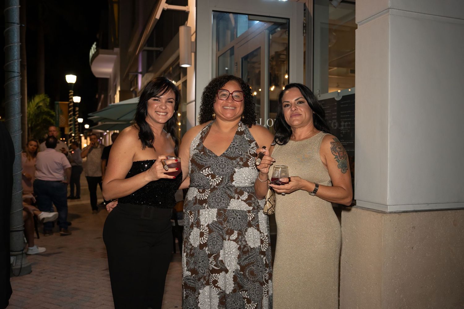 Three women standing together with drinks outside during a grand opening event.