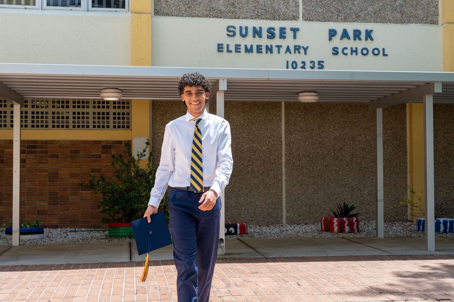 Young graduate stands in front of Sunset Park Elementary School, dressed in tie and cap for milestone portraits in Miami, Florida.