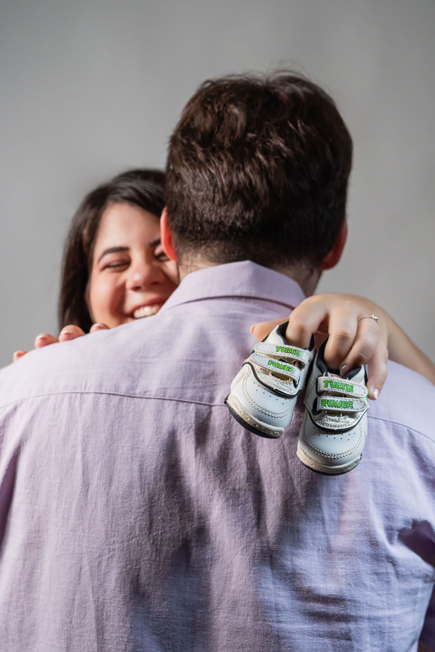 Maternity studio photo of woman holding baby shoes behind partner’s back
