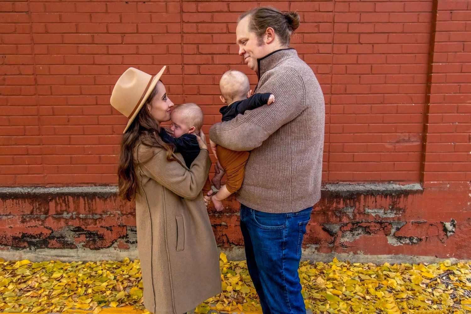Romantic fall couple photoshoot in Denver, CO – parents holding their newborns against a red brick wall with golden leaves.