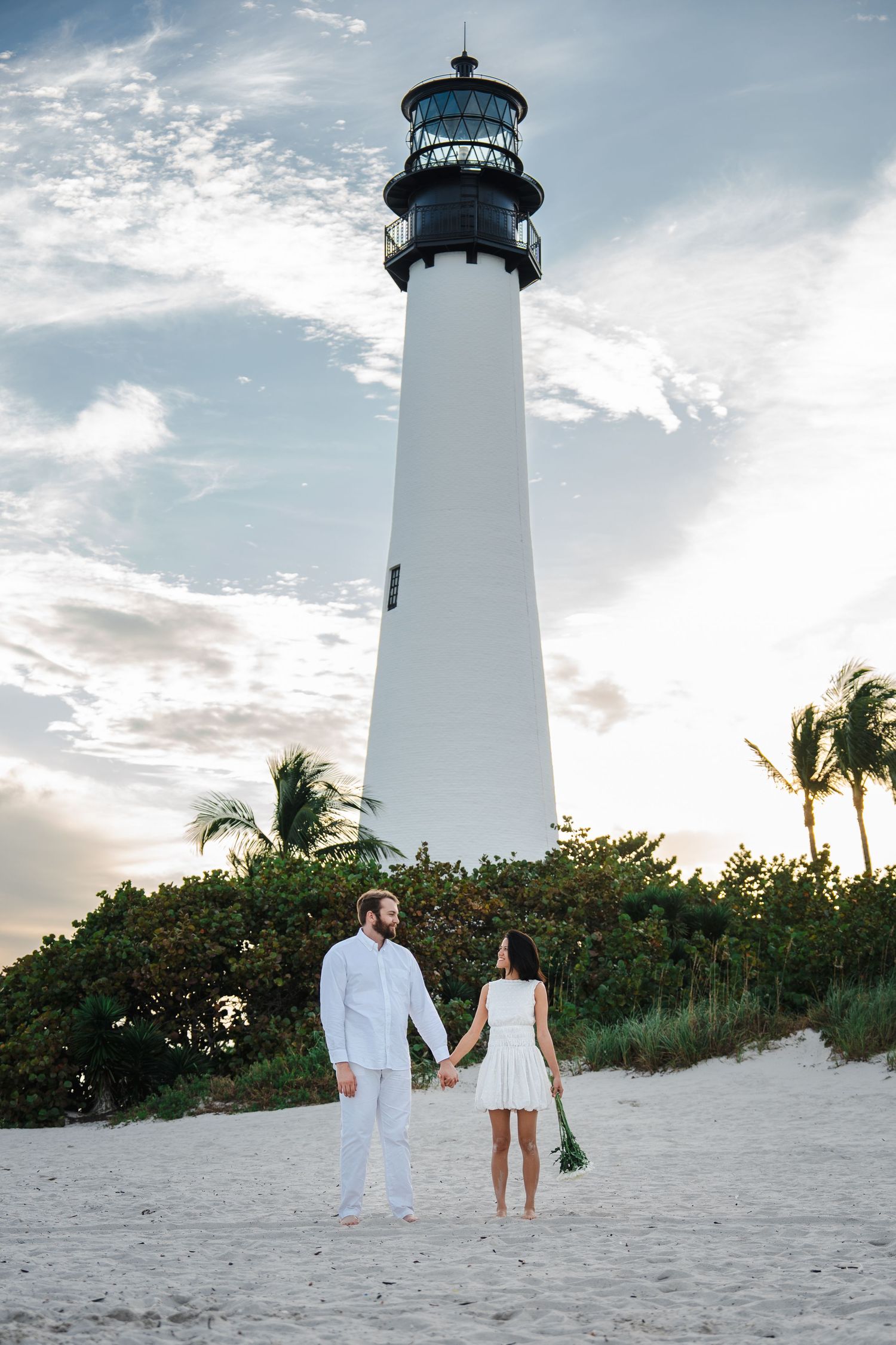 Couple holding hands beneath the Cape Florida lighthouse at Bill Baggs State Park during a romantic Miami engagement session.