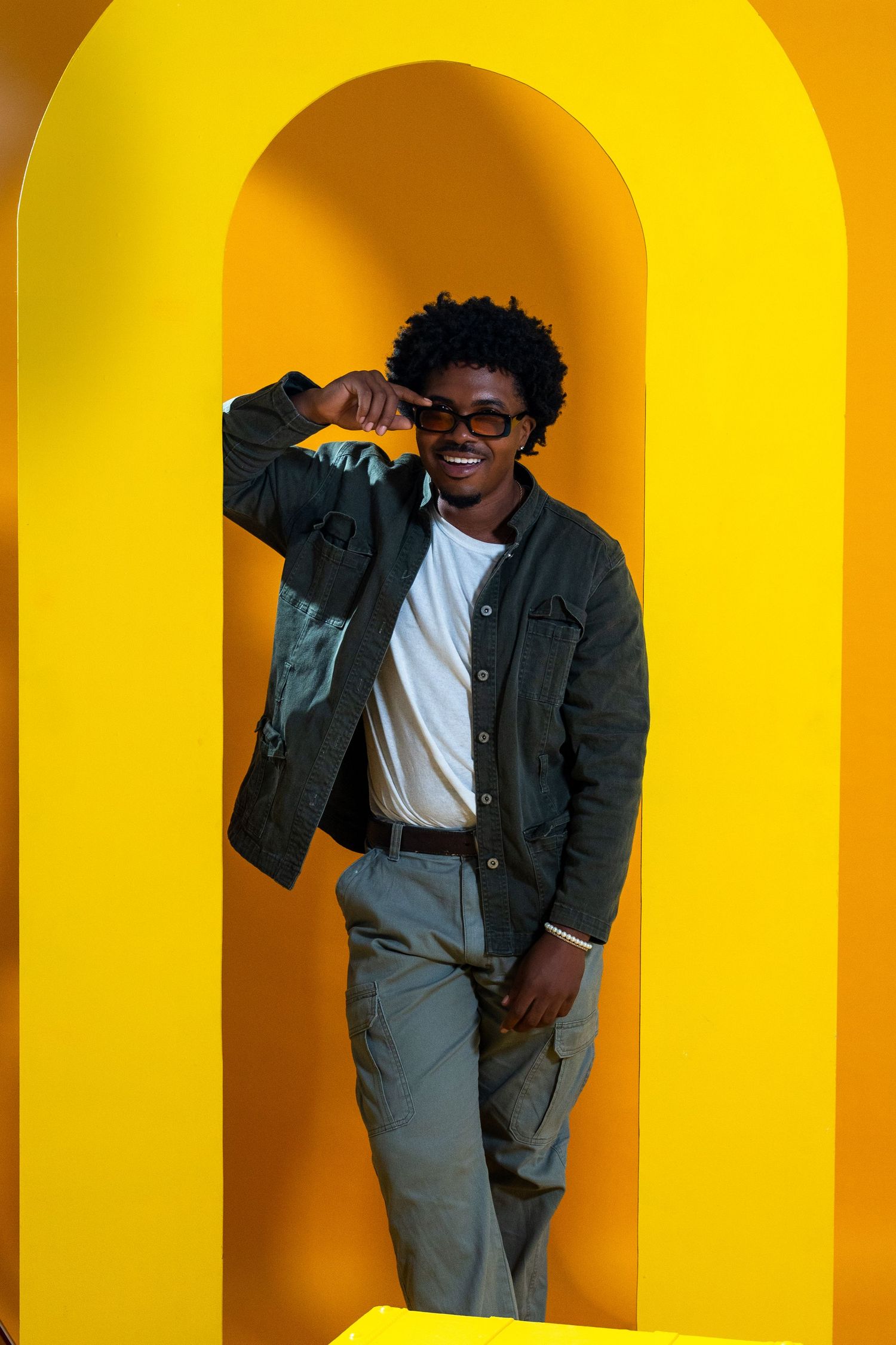 Modern editorial portrait in South Florida – male model in a green jacket and glasses walks through a bold yellow archway.