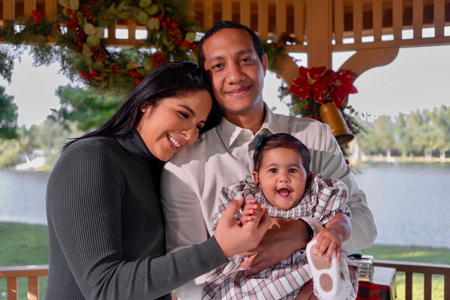 Joyful holiday family portrait in Fort Lauderdale, FL – parents holding their baby with a Christmas backdrop by the lake.