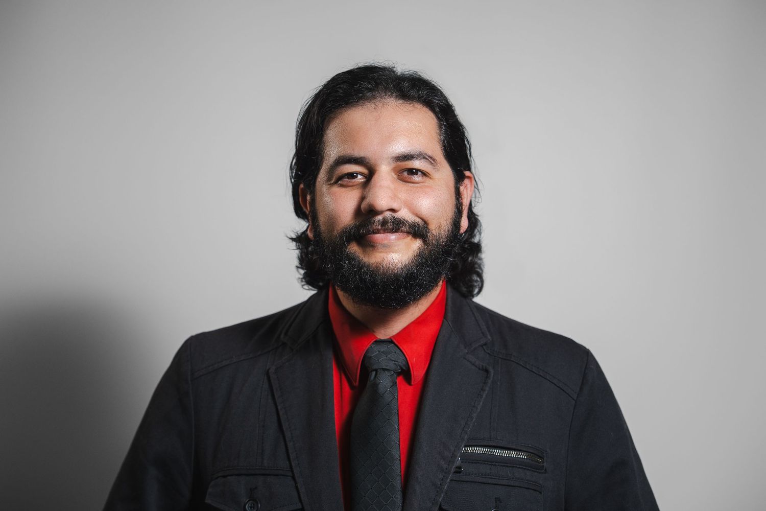 Man in black suit with red shirt and tie smiling against gray studio background