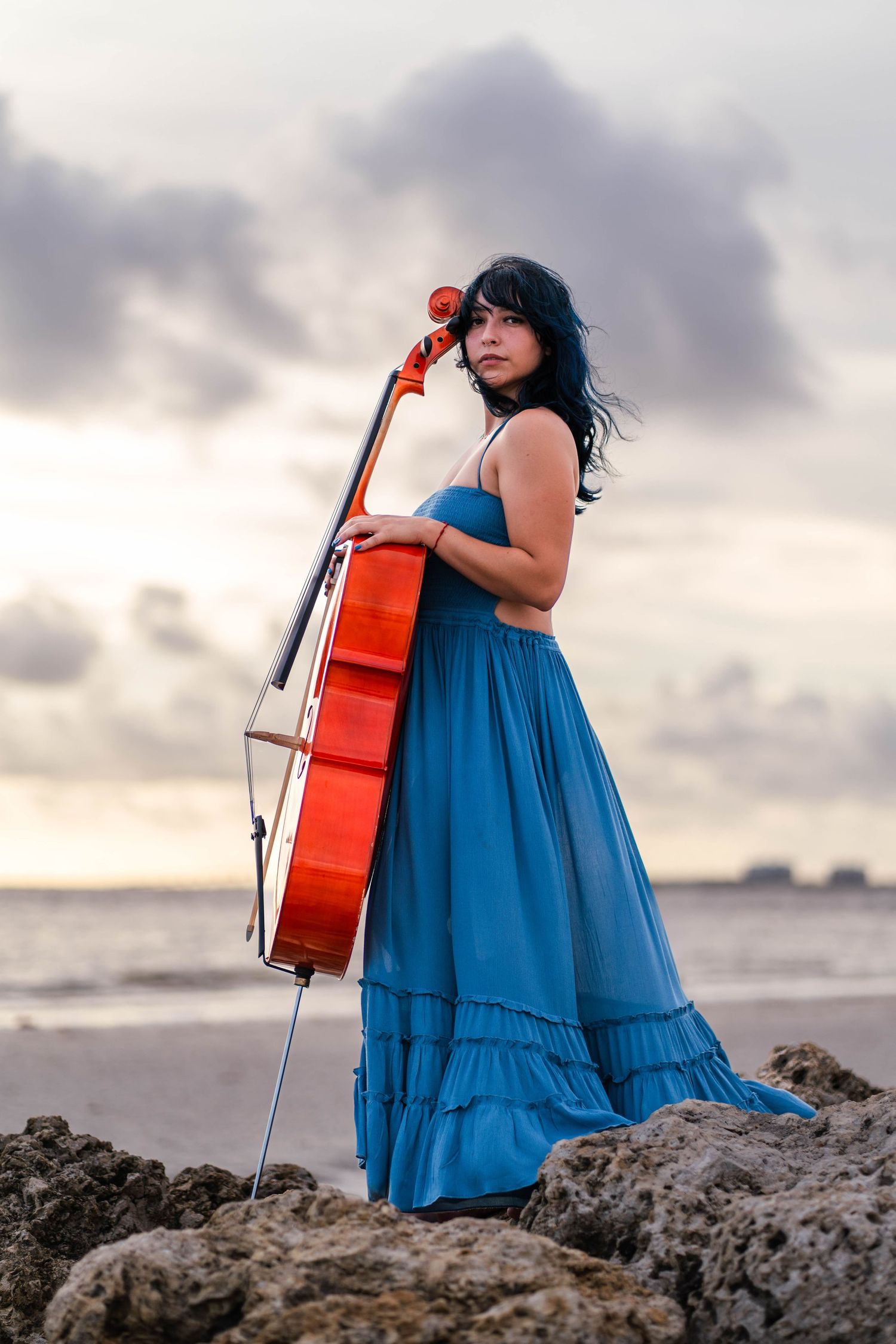Cellist in blue dress posing with cello on rocks during South Florida portrait session