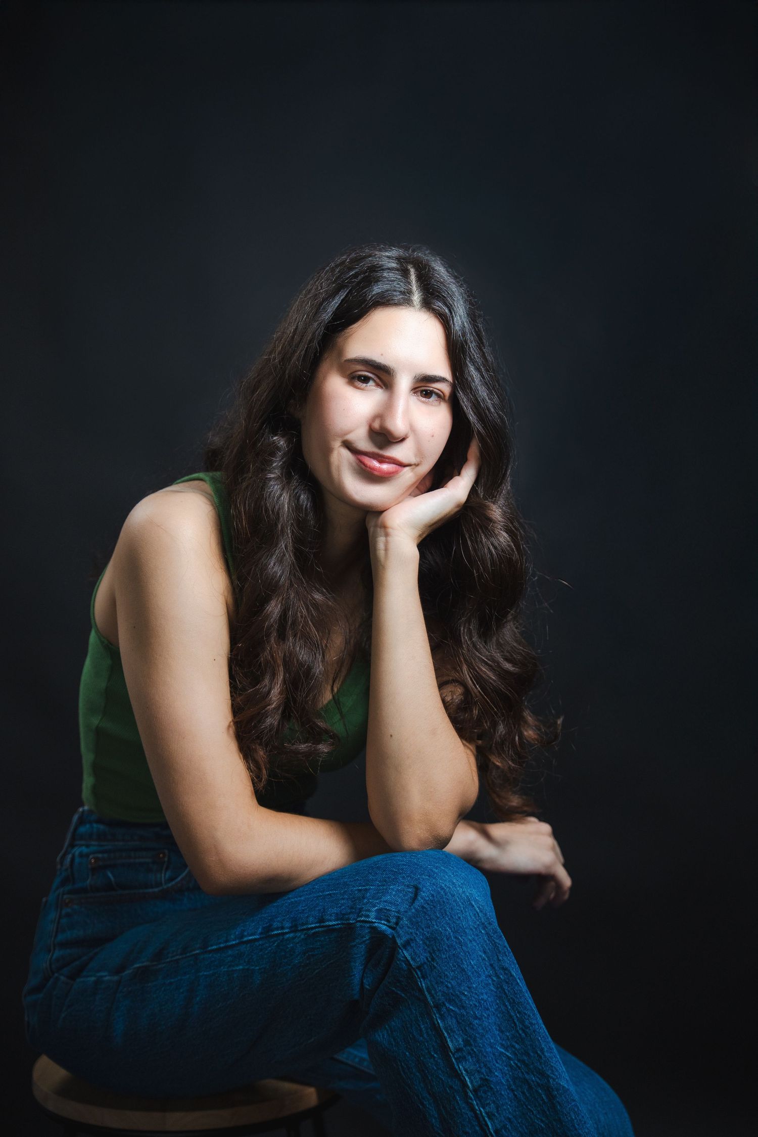 Studio acting portrait of a woman seated with her chin resting on her hand, photographed against a dark backdrop.
