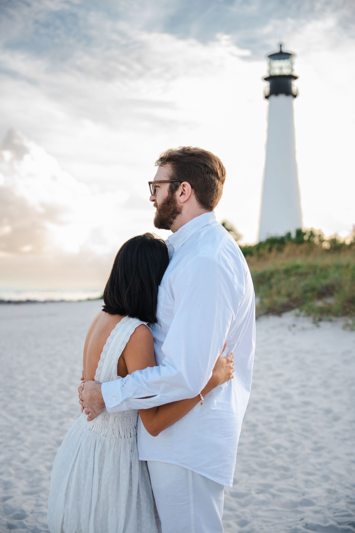 Engaged couple standing together on the beach with the Cape Florida lighthouse in the background at sunset.