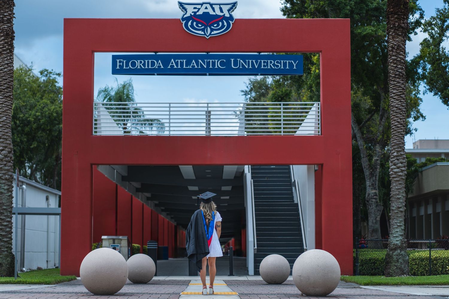 Florida Atlantic University graduate in cap and gown walking toward FAU sign, celebrating her achievement in Boca Raton, Florida.