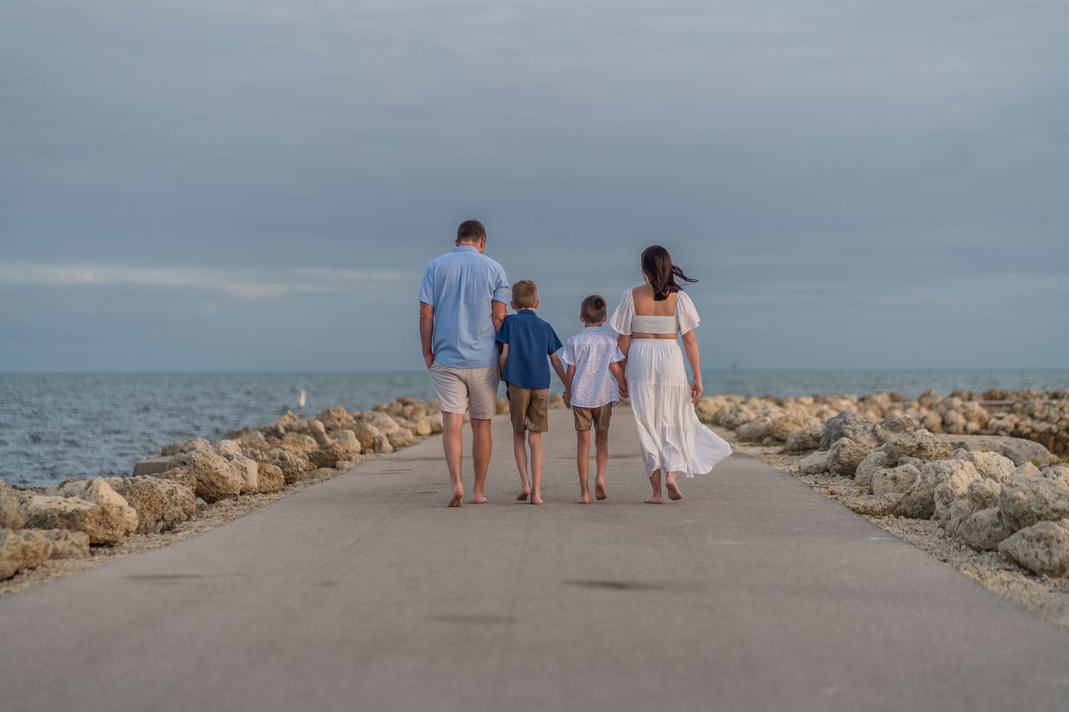 Coastal family portrait in Harry Harris Park, FL – family walking hand in hand along a scenic rocky pier with an ocean backdrop.