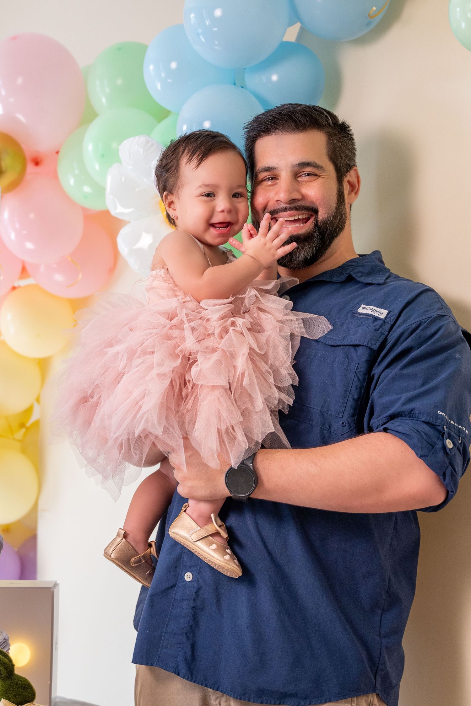 Father smiling while holding his baby daughter in a pink dress during an indoor first birthday photo session.