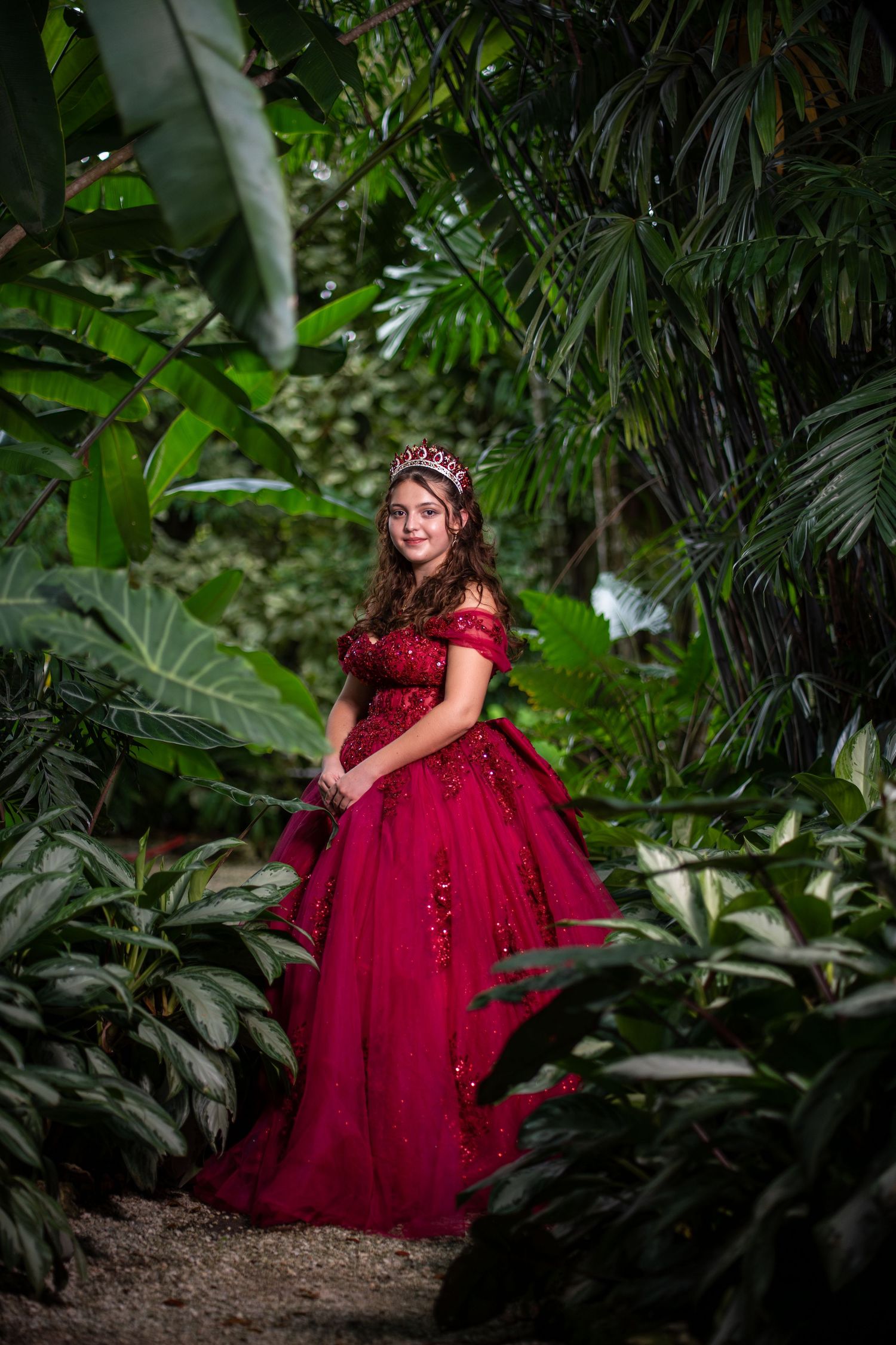 Quinceañera portrait in a flowing burgundy ball gown posed in front of a garden waterfall.