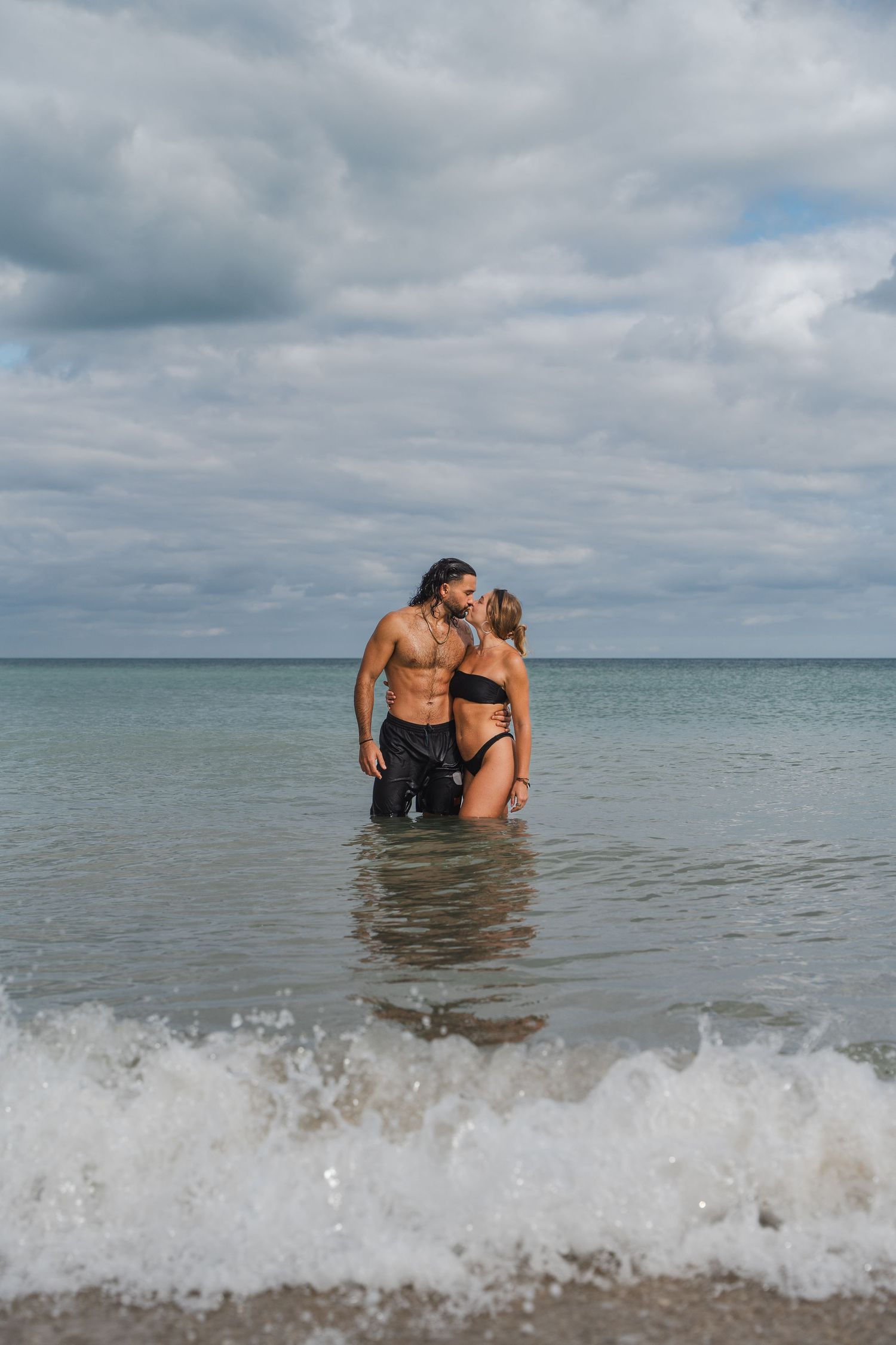 Wide beach portrait of a couple standing together in shallow water with waves and an overcast sky behind them.