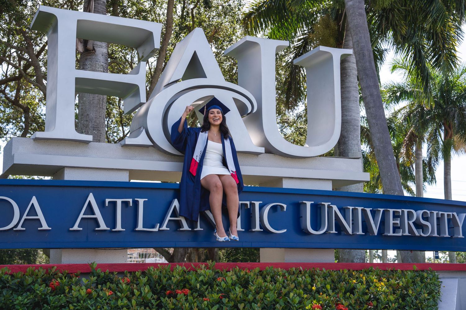FAU graduate posing by university sign in Boca Raton wearing cap and gown