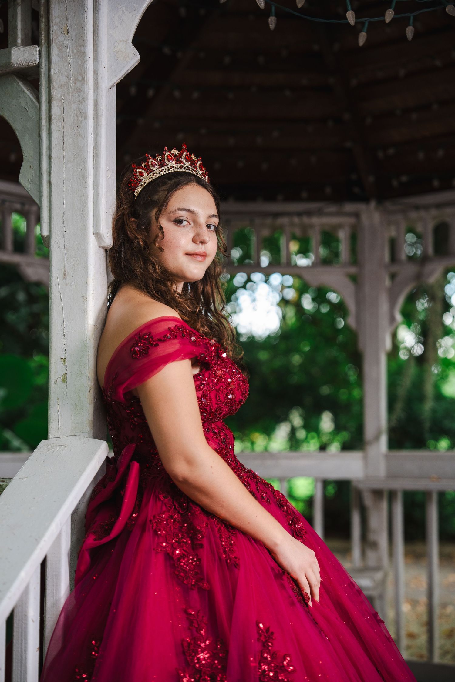Quinceañera portrait in a burgundy gown and tiara, posed under a gazebo at a lush orchid garden.
