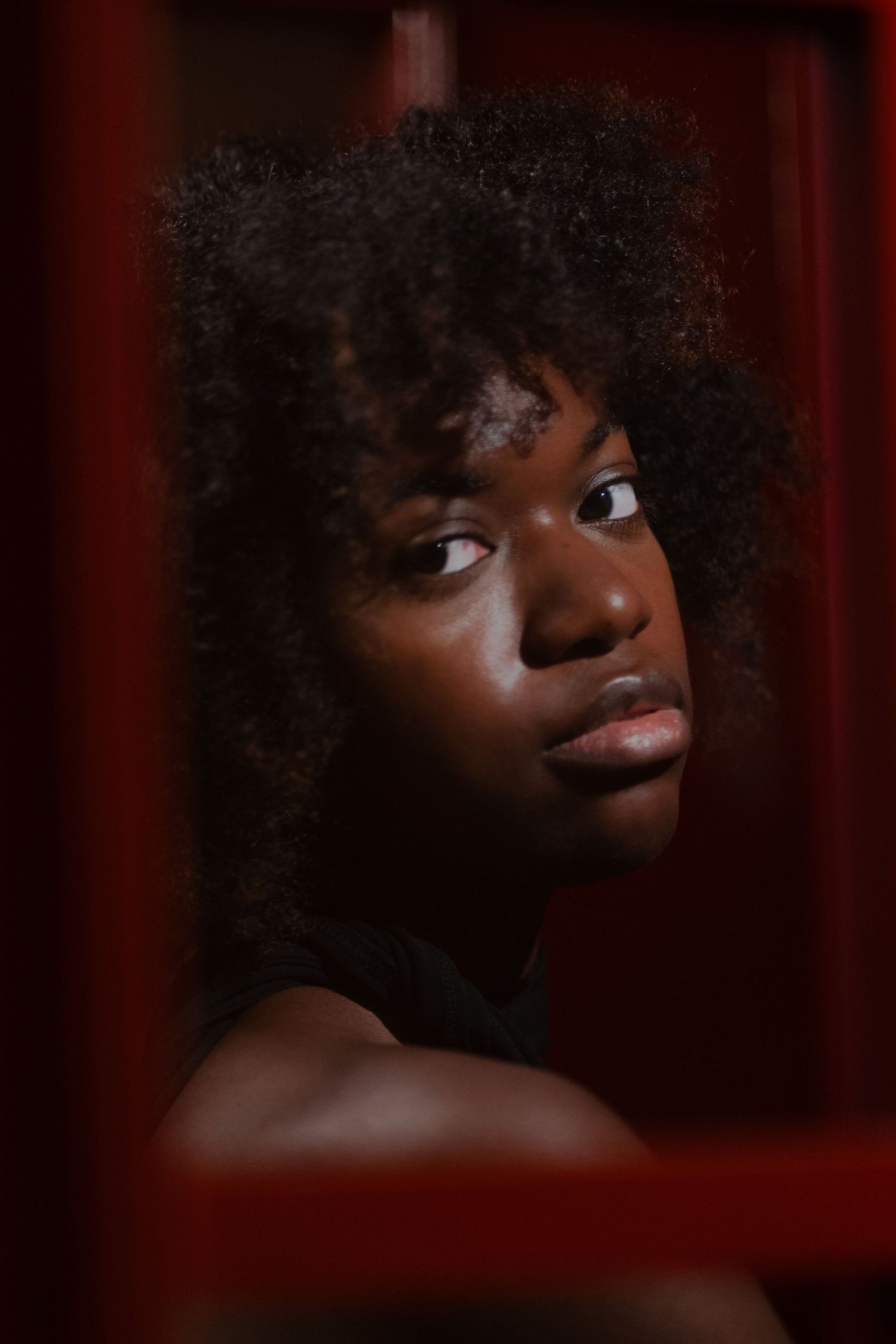 Dramatic editorial portrait at a private studio – woman with natural curls gazes through a red-framed doorway, creating an artistic moody vi