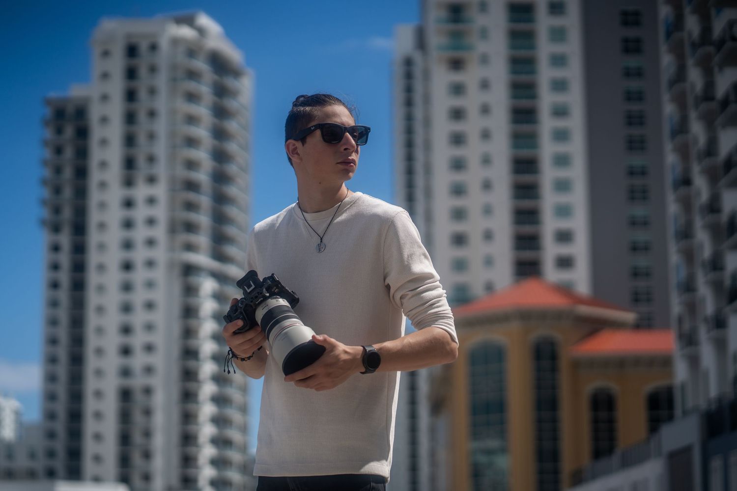 Photographer in light sweater holding camera in downtown Fort Lauderdale with high-rise buildings in the background.
