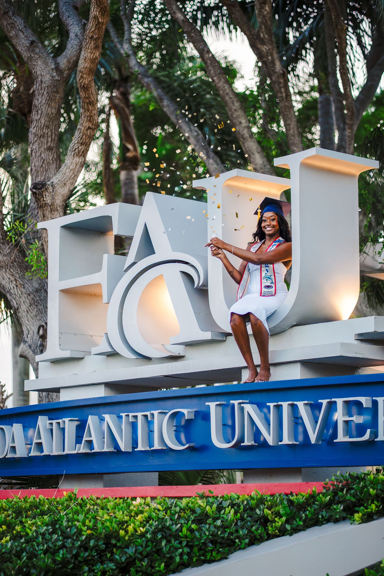 Guided FAU graduation portrait session with graduate posing on Florida Atlantic University sign in Boca Raton popping confetti