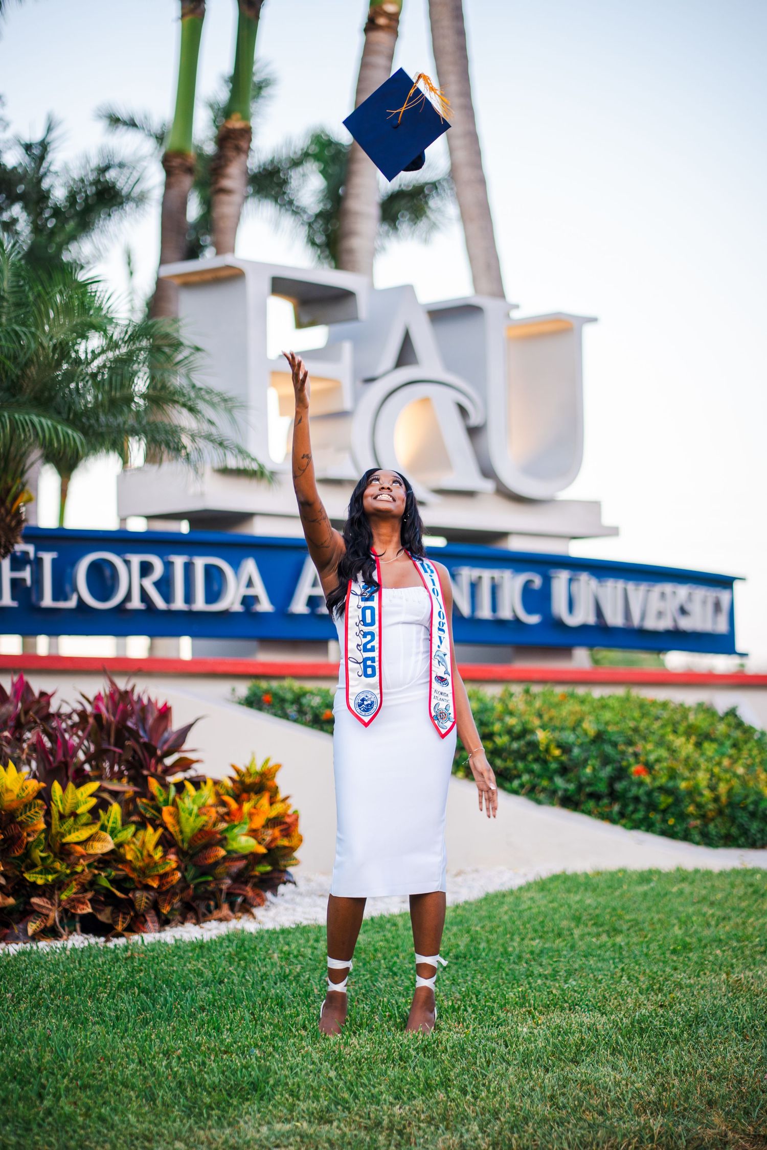 Graduate in white dress tosses cap at Florida Atlantic University sign, celebrating her FAU graduation outdoors.